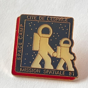 Op de afbeelding: Een rechthoekige speld met een rode rand en gouden afwerking. De speld heeft een donkerblauwe achtergrond met een gouden astronautenillustratie en de tekst "CITE DE L'ESPACE", "SPACE CAMP" en "MISSION SPATIALE 91".