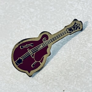 May include: A vintage-style mandolin pin with a maroon body and a tan border. The pin features black details for the strings and fretboard. The headstock has a decorative design.
