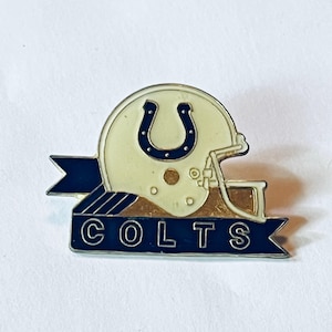 May include: A metal enamel pin featuring an off-white football helmet with a blue horseshoe logo. A dark blue banner sits across the bottom with the word COLTS in silver capital letters. The design includes a silver face mask and metallic accents.