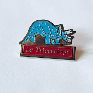May include: A metal enamel pin featuring a bright blue triceratops dinosaur silhouette above a rectangular red banner with a gold border. The text Le Triceratops is printed in gold lettering inside the red banner against a white background.