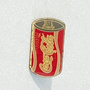 May include: A rectangular pin featuring a red Coca-Cola can design. The pin has a gold border and the Coca-Cola logo in gold script. The top of the can is gray with white accents. The pin is likely made of metal.