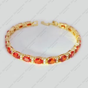 以下が含まれることがあります： 楕円形の赤い宝石と小さな透明な宝石が並んだ金のブレスレット。