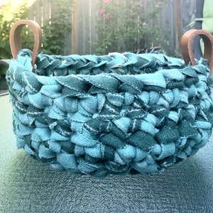 Peut inclure: Un panier rond en tissu turquoise avec des poignées en cuir marron. Le panier est fait de bandes de tissu tressées, créant un aspect texturé. Les poignées sont fixées sur le bord supérieur, offrant un élément décoratif et fonctionnel.