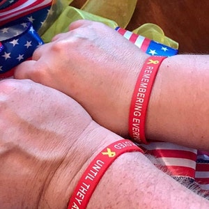 May include: Two red wristbands with yellow ribbons and white text. The text on the left wristband reads "NEVER UNTIL THEY ARE LOVED". The text on the right wristband reads "REMEMBERING EVERY LOVED".