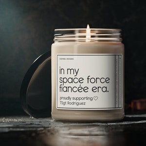 May include: A beige candle with a white label that reads "in my space force fiancée era. proudly supporting♡ TSgt Rodriguez".