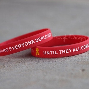 May include: Two red silicone bracelets with white text. The top bracelet reads "Remembering Everyone Deployed". The bottom bracelet reads "Until They All Come Home" and has a yellow ribbon graphic.