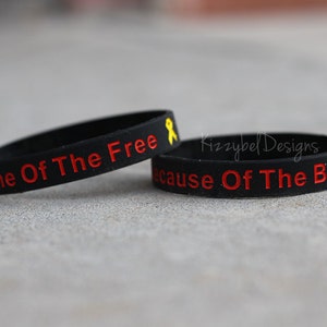 May include: Two black silicone bracelets with red text. The top bracelet reads "Home Of The Free" and has a yellow ribbon. The bottom bracelet reads "Because Of The Brave".