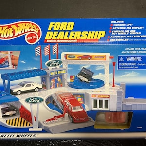 May include: A Hot Wheels Ford Dealership playset in a blue box. The playset includes a showroom, sales office, and car wash. The box features the Hot Wheels logo and the Ford logo. The playset includes a working lift and a rotating car display.