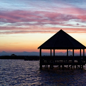 May include: Silhouette of a wooden gazebo on a pier over a body of water at sunset. The sky is a vibrant mix of pink, orange, and purple.