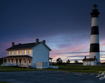 May include: A white lighthouse with black stripes stands tall against a twilight sky. A smaller white house with windows lit up is in the foreground. The sky is a mix of purple, pink, and blue.