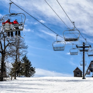 May include: Two people ride a ski lift up a snowy mountain on a sunny day. The lift is full of empty chairs and the sky is blue with white clouds.