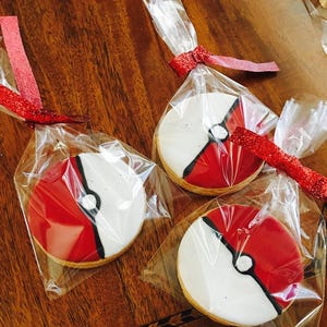 May include: Three individually packaged cookies decorated with red, white, and black icing to resemble a Pokeball. The cookies are wrapped in clear plastic bags with red ribbon.