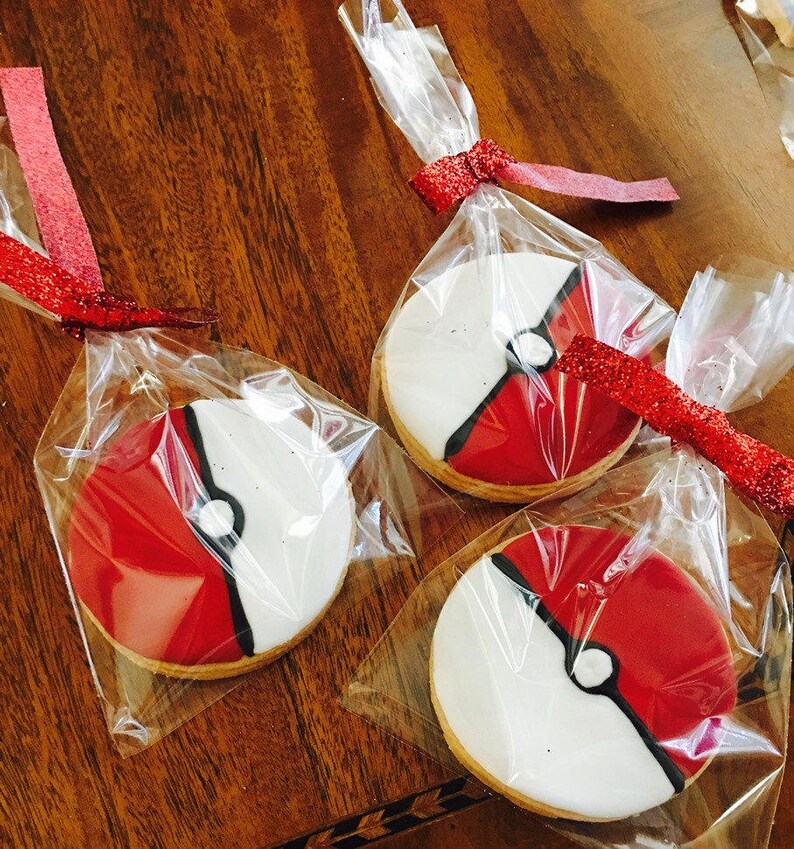 May include: Three individually packaged cookies decorated with red, white, and black icing to resemble a Pokeball. The cookies are wrapped in clear plastic bags with red ribbon.