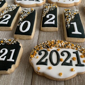 May include: A close-up of nine decorated sugar cookies with black and white icing. The cookies are decorated with the year 2021 and gold and white sprinkles.