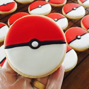 May include: A red, white, and black cookie decorated with a Pokeball design. The cookie is held in a gloved hand.