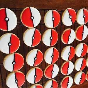 May include: A close-up of a cooling rack with 24 round cookies decorated with red, white, and black icing. The cookies are arranged in rows and are all the same size and shape. The cookies are decorated with a design that resembles a Pokeball.