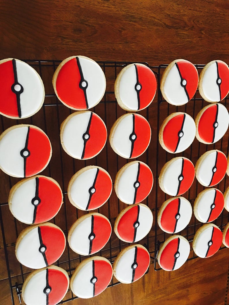 May include: A close-up of a cooling rack with 24 round cookies decorated with red, white, and black icing. The cookies are arranged in rows and are all the same size and shape. The cookies are decorated with a design that resembles a Pokeball.