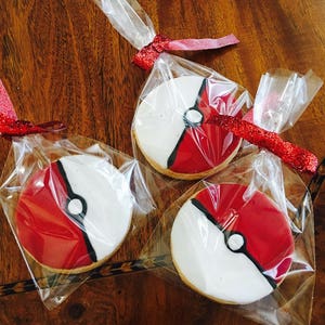 May include: Three individually packaged cookies decorated with red, white, and black icing to resemble a Pokeball. The cookies are packaged in clear plastic bags tied with red ribbon.