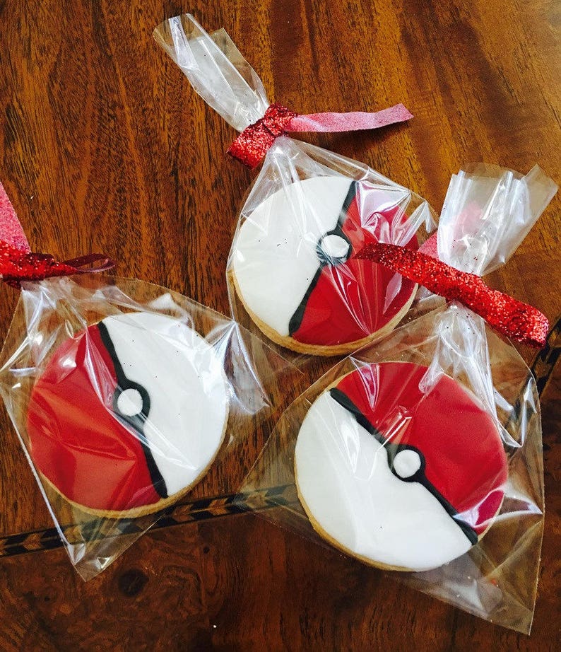May include: Three individually packaged cookies decorated with red, white, and black icing to resemble a Pokeball. The cookies are packaged in clear plastic bags tied with red ribbon.