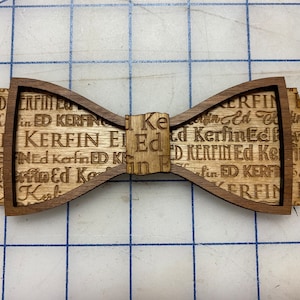 May include: A wooden bow tie with a laser-engraved pattern of the name "Ed Kerfin" repeated throughout the design.