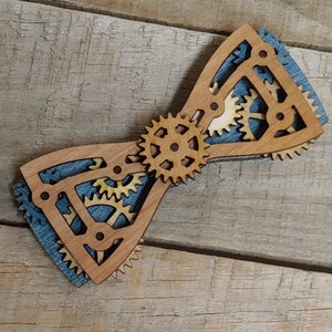 May include: A wooden bow tie with a blue and beige patterned design featuring intricate gear cutouts. The bow tie is made of two pieces of wood that are joined together in the centre.