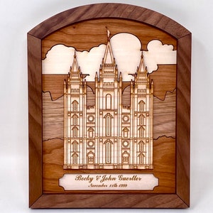 Puede incluir: Una pieza de arte de pared enmarcada en madera con un diseño de templo cortado con láser con una nube blanca encima. El marco es de madera marrón oscuro con una parte superior arqueada. El texto "Becky & John Guertler, November 11th 1999" está grabado en una placa debajo del templo.