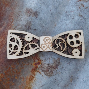 Può includere: Un papillon in legno con un design steampunk che presenta intricati ritagli di ingranaggi e pignoni. Il papillon è realizzato in legno marrone chiaro con un centro in legno marrone più scuro.