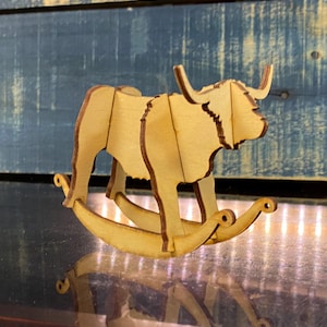 Puede incluir: Un juguete de madera para mecer en forma de vaca de las tierras altas. El juguete está hecho de madera marrón claro y tiene un diseño simple. La vaca tiene una melena larga y fluida y una cola larga.