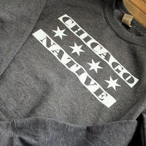 May include: A gray sweatshirt with a white graphic design featuring the words "CHICAGO NATIVE" and four white stars.
