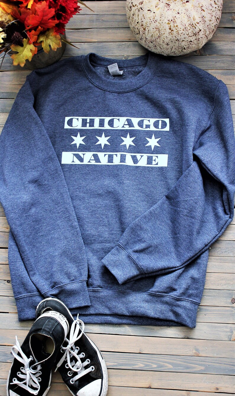 May include: A dark blue sweatshirt with white text that reads "CHICAGO*****NATIVE". The sweatshirt is laying on a wooden surface with a pair of black and white sneakers.