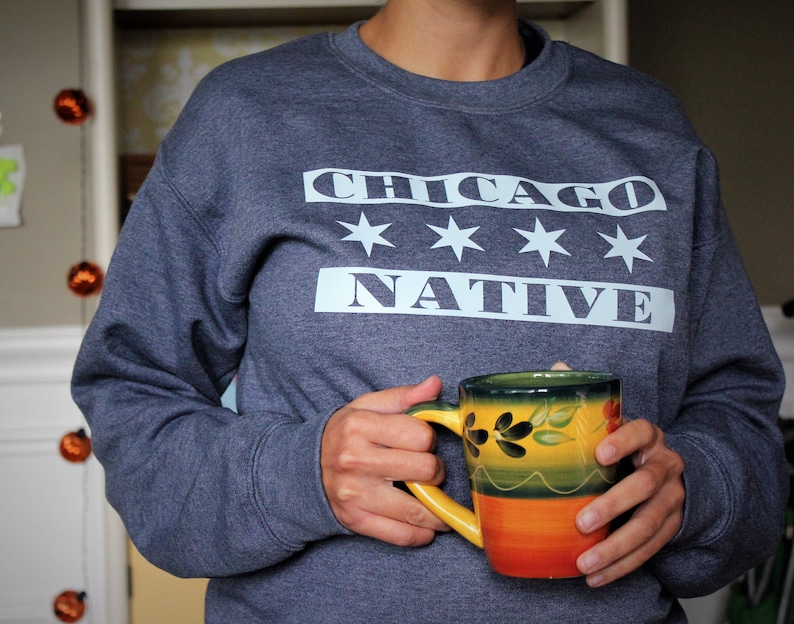 May include: A dark blue sweatshirt with the text "CHICAGO NATIVE" in white letters on the front. The sweatshirt has a white star design above the text. A person is holding a yellow and green ceramic mug.