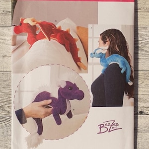 May include: Simplicity sewing pattern featuring plush dragon and horse designs. The pattern includes a red dragon, a blue dragon, and a purple horse. The pattern is labeled "8715 OS" and includes the text "Bee Zee Art" and "Since 1927".