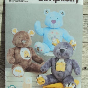 May include: A Simplicity sewing pattern for making three plush teddy bears. The pattern includes instructions for making a blue teddy bear, a brown teddy bear, and a gray teddy bear. Each bear has a unique design with embroidered details. The pattern is labeled S9731 and is for one size.