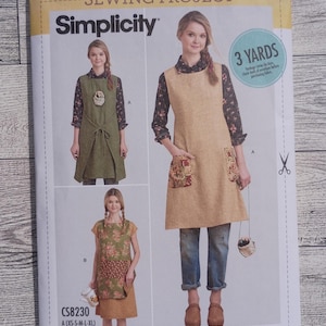 May include: A Simplicity sewing pattern envelope for apparel project CS8230 by Dottie Angel. The cover displays three variations of sleeveless tunics or aprons in olive green and tan, featuring floral pocket details, tie-waist options, and layered fabric designs.