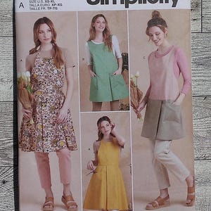 May include: A sewing pattern for a dress with an apron overlay. The pattern includes three different styles: a floral print dress with a brown apron, a green dress with a green apron, and a pink dress with a beige apron. The pattern is labeled "S9564" and "Simplicity".