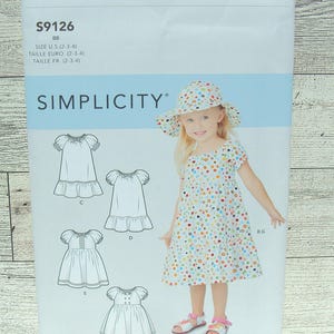 May include: A Simplicity sewing pattern for a girl's dress, sizes 2-3-4. The pattern includes five different styles of dresses, all with a gathered neckline and short sleeves. The pattern also includes instructions for making a matching sun hat.