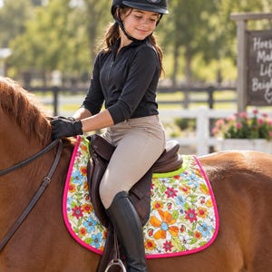 May include: A quilted horse saddle pad featuring a vibrant floral pattern with orange, red, and blue flowers on a cream background, finished with a bright pink trim. The pad is positioned under a brown leather saddle on a chestnut horse.