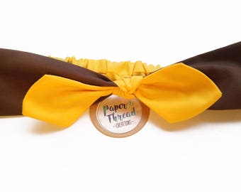 May include: A brown and yellow fabric headband with a bow. The headband has a small tag that says "Paper & Thread - Creations".