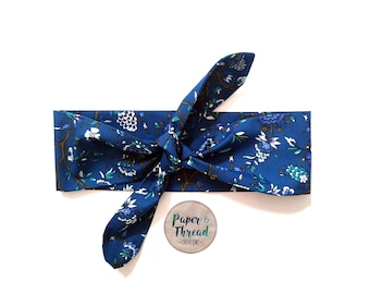 May include: A blue headband with a floral pattern. The headband is tied in a knot at the top. The text "Paper & Thread - Creations" is visible on a tag.