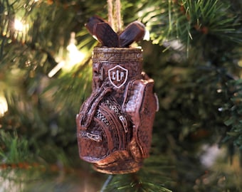 Puede incluir: Adorno navideño de bolsa de golf marrón con palos de golf. El adorno tiene una apariencia texturizada y un emblema blanco con las letras "CLP". El adorno está colgado en un árbol de Navidad.