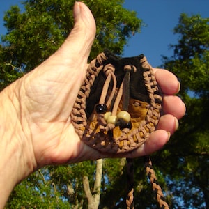 May include: A small, handmade pouch with a woven brown border and a black interior. The pouch has a drawstring closure and is decorated with beads. The background features green trees and a blue sky.