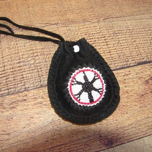 May include: Black beaded pouch with a drawstring closure and a long black cord. The pouch features a circular design with a white and red border, and a black starburst pattern in the center. The pouch is displayed on a wooden surface.