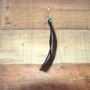May include: A keychain with a silver clasp and chain, adorned with a small charm and a dark brown tassel. The tassel is made of multiple strands of hair-like material, and a small green bead is visible.
