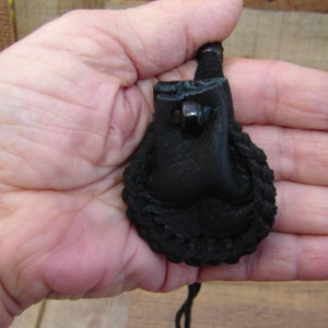 May include: A small, black leather pouch with a braided edge and a drawstring closure. The pouch is held in a hand, showcasing its compact size and detailed craftsmanship. The leather appears to be textured, adding to its rustic appeal.