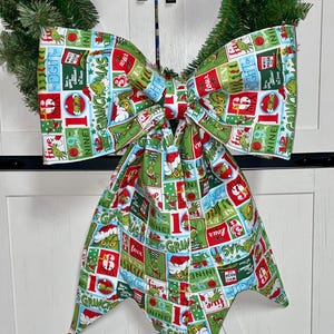 May include: A large, decorative bow made of fabric featuring a colorful Grinch-themed pattern. The bow is predominantly green, red, and white, with various Grinch illustrations and text. The bow is attached to a green wreath, suitable for holiday decor.
