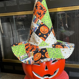 May include: A pointed witch hat featuring a patchwork design of Halloween-themed fabric. The pattern includes orange pumpkins, black cats, candy corn, and colorful book spines on white, black, and lime green backgrounds with star accents.