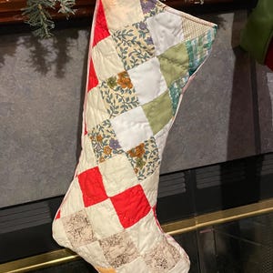 May include: A quilted Christmas stocking with a patchwork design. The stocking features squares of red, white, and patterned fabrics in shades of green, beige, and orange. The stocking is hanging near a fireplace and Christmas tree.