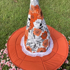 Puede incluir: Un sombrero de bruja de Halloween con un ala ancha naranja y una corona alta y puntiaguda. La corona está decorada con rosas naranjas, detalles grises y telarañas sobre un fondo blanco. Un ribete de encaje blanco bordea la corona.