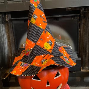 Puede incluir: Un sombrero de bruja puntiagudo con un diseño de patchwork de tela naranja con mapaches y tela a cuadros negros y grises. El sombrero descansa sobre una calabaza de Halloween naranja con ojos triangulares y una boca ancha tallada.