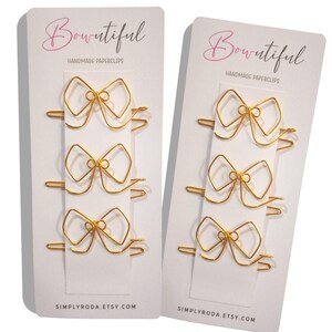 Op de afbeelding: Twee witte kaarten met gouden strikvormige paperclips. Op elke kaart staat de tekst "Bowtiful Handmade Paperclips" en de website "simplyroda.etsy.com".